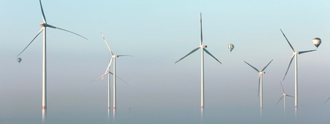 Luftaufnahme von Windkraftanlagen mit einem Heissluftballon, der in der Nähe schwebt, im sanften Morgenlicht.