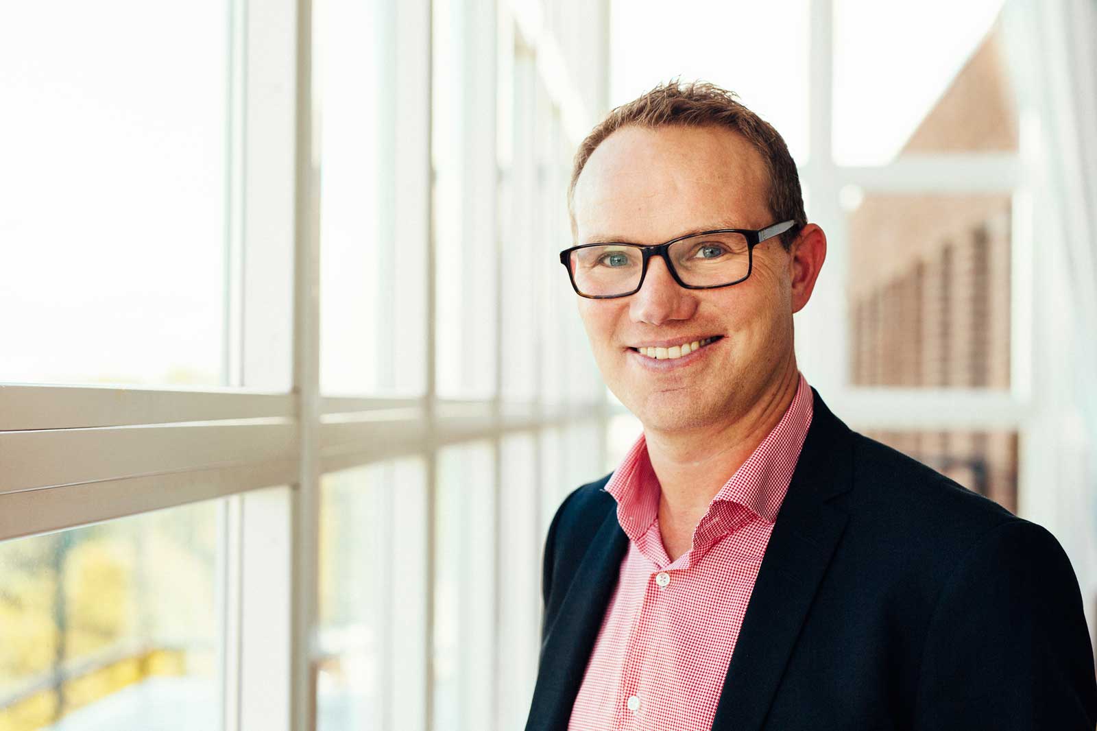 A professionally dressed individual stands beside large windows, with natural light illuminating the scene.