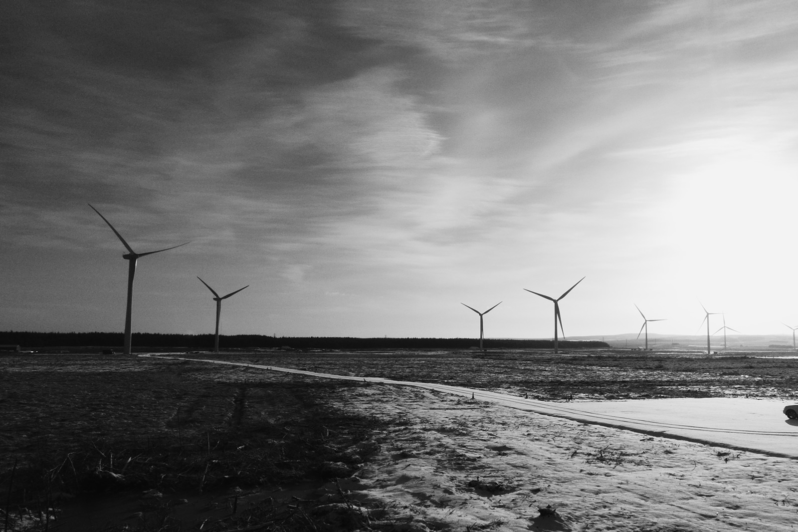A black and white landscape featuring multiple wind turbines against a cloudy sky, with a path running through a field.