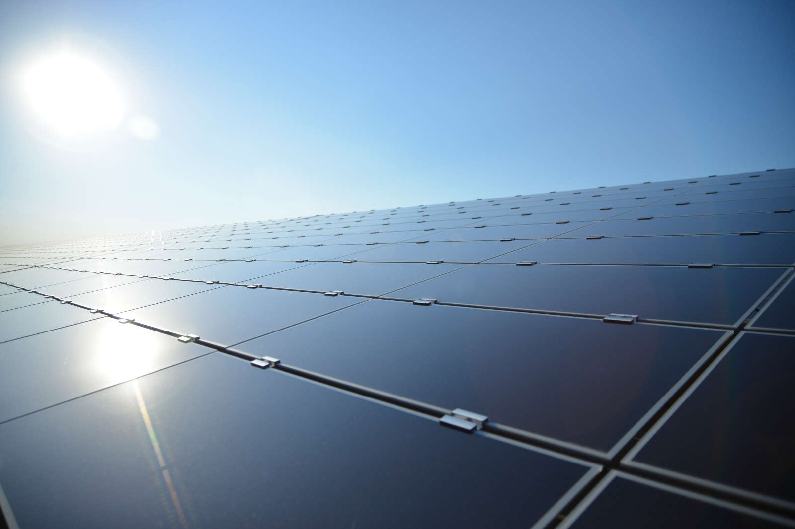 A solar panel field with glossy panels against a clear blue sky and the sun shining in the background.