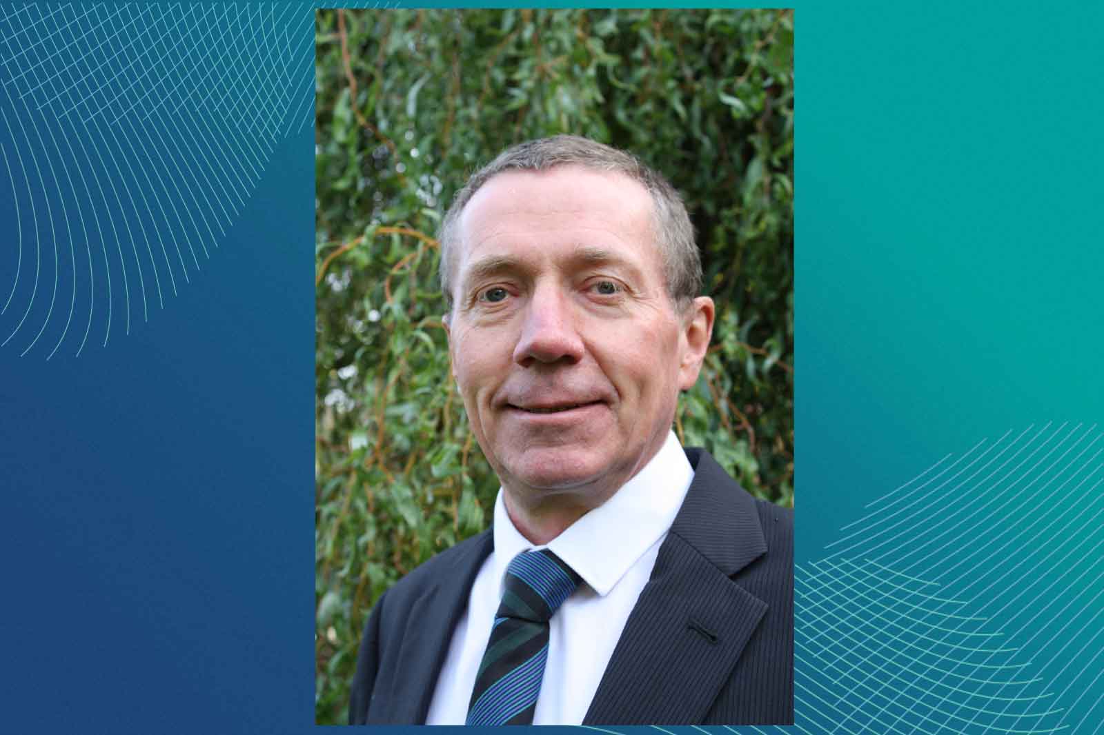 A man in a black suit and blue tie stands outdoors against a background of greenery and abstract blue patterns.