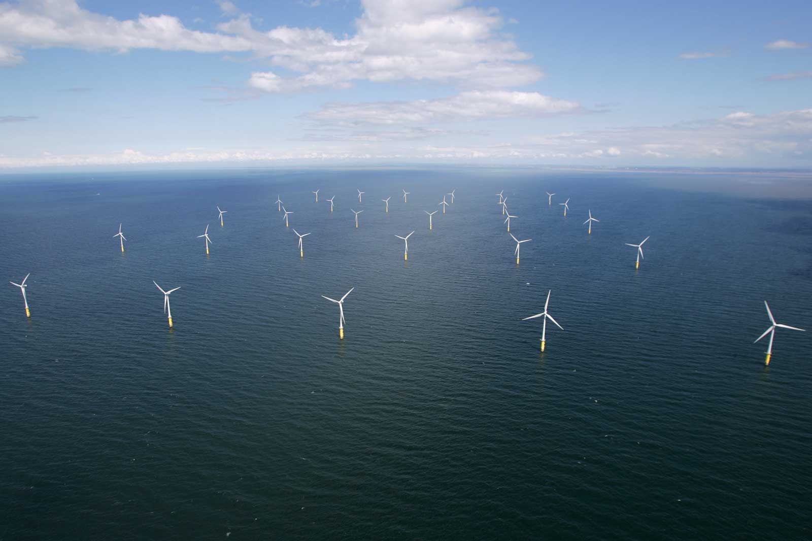 An offshore wind farm featuring numerous wind turbines standing in a vast ocean, under a blue sky with some clouds.