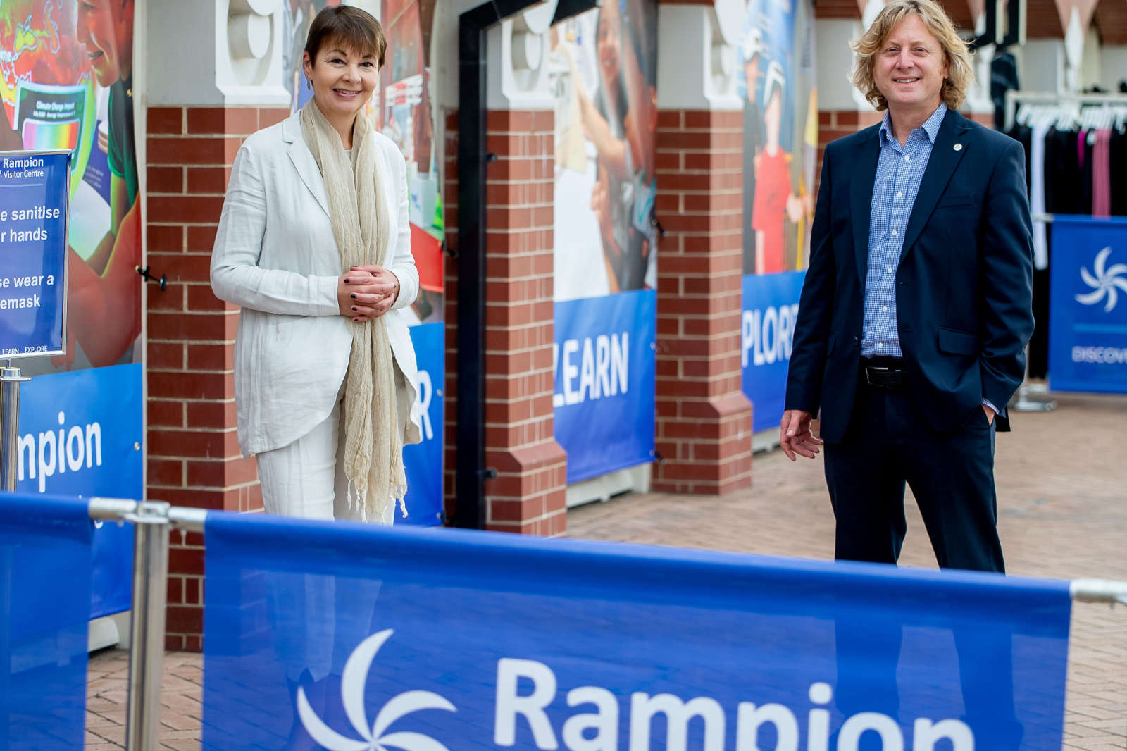 A woman in a light outfit and a man in a suit stand in front of banners promoting safety measures for visitors.