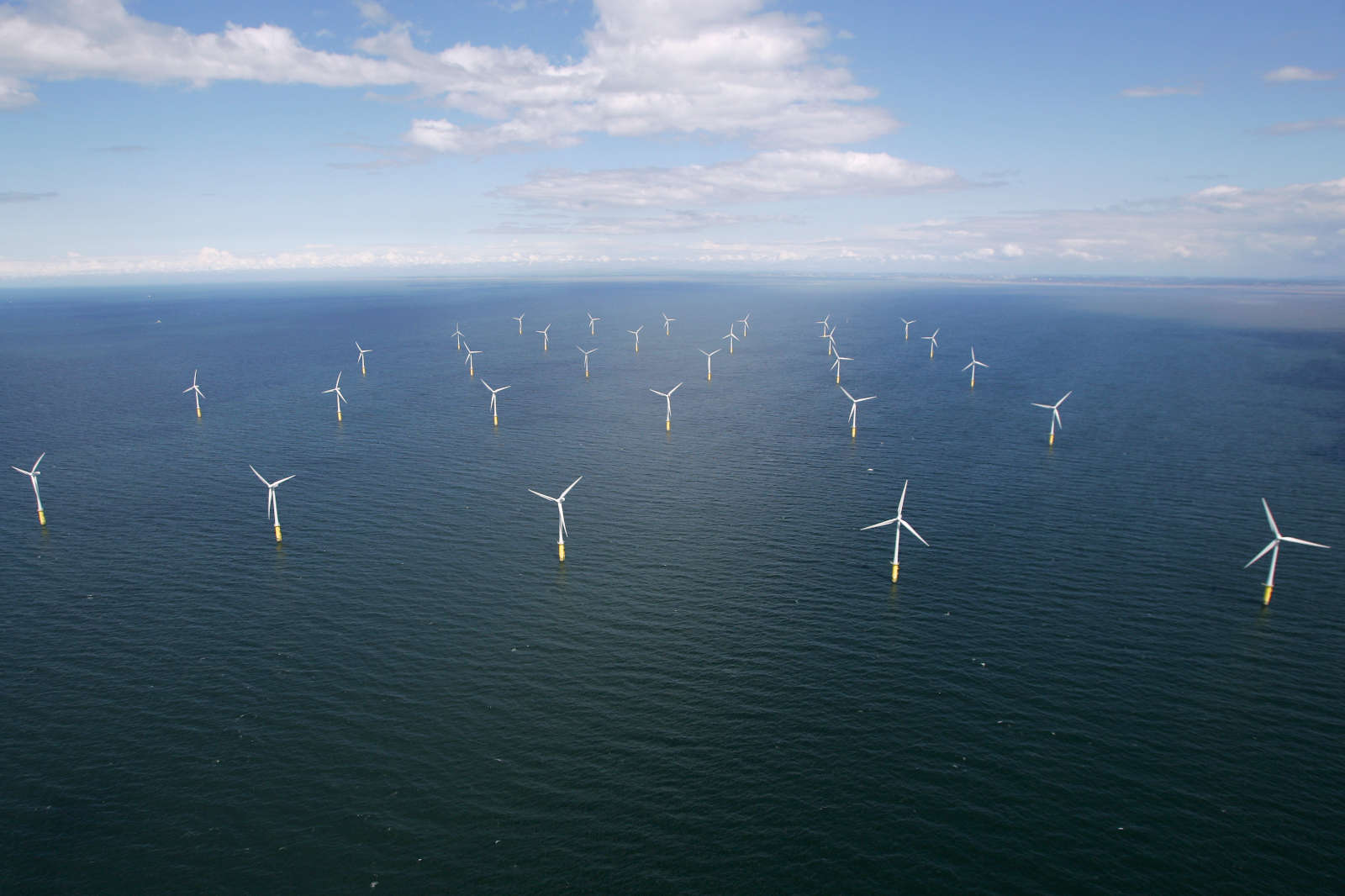 An aerial view of an offshore wind farm featuring numerous wind turbines on the water beneath a blue sky.