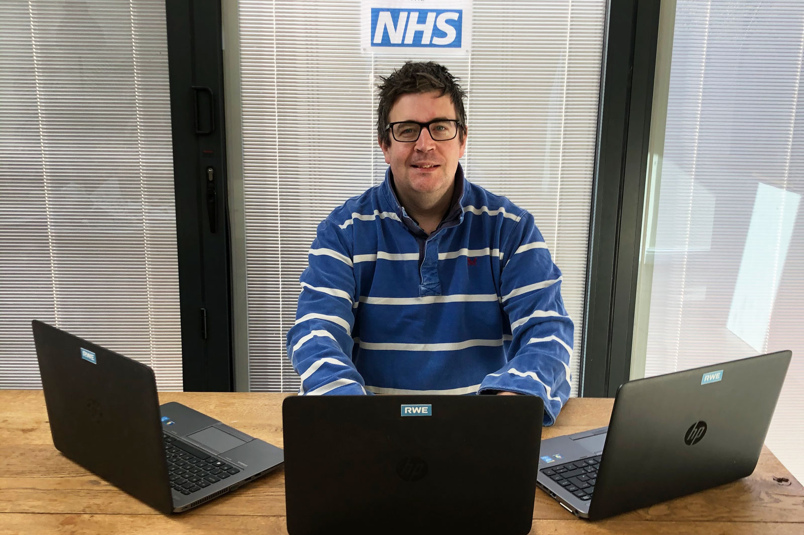 A person wearing a blue and white striped shirt sits at a wooden table with three laptops open, behind an NHS sign.