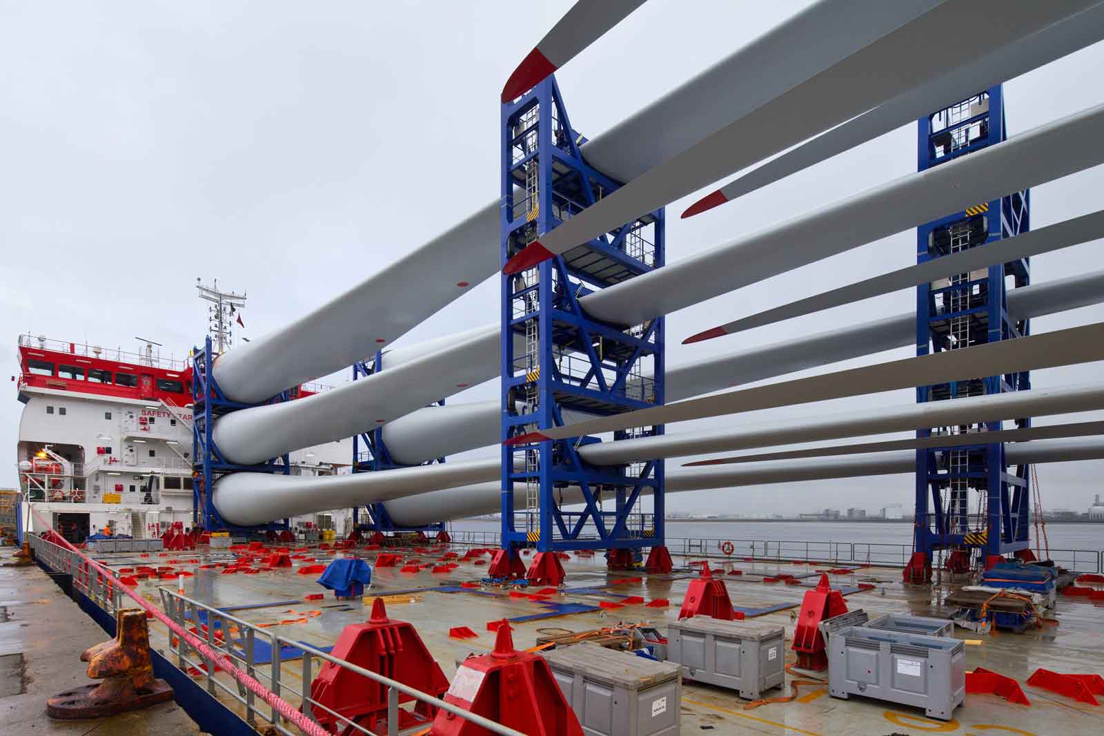 A shipping vessel with large wind turbine blades stacked vertically on a deck. The scene is set on a cloudy day, showing machinery.