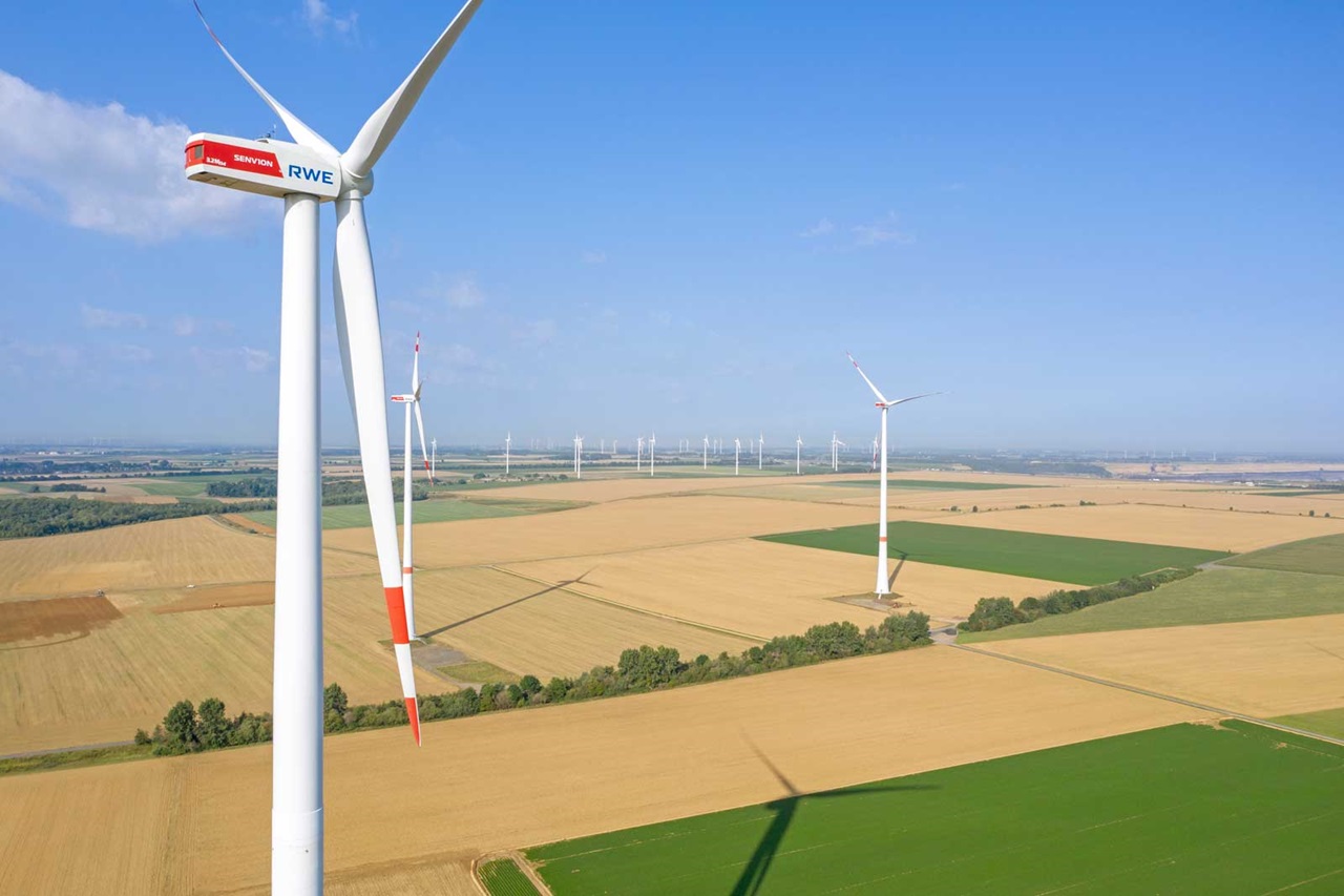 Luftaufnahme von Windkraftanlagen in einer weiten Landwirtschaftslandschaft unter blauem Himmel.