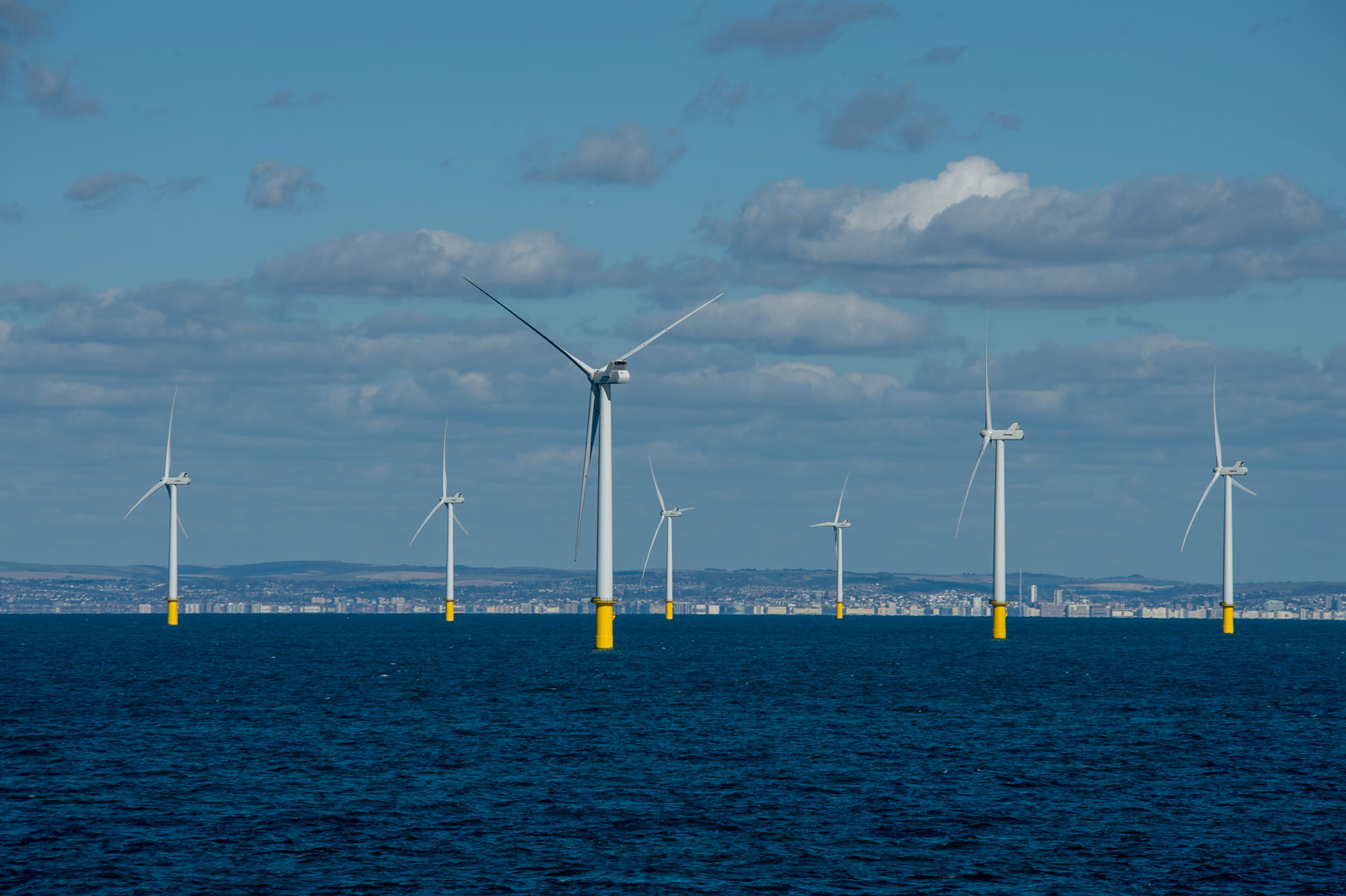 A row of offshore wind turbines with yellow bases stands in the sea under a blue sky with scattered clouds.