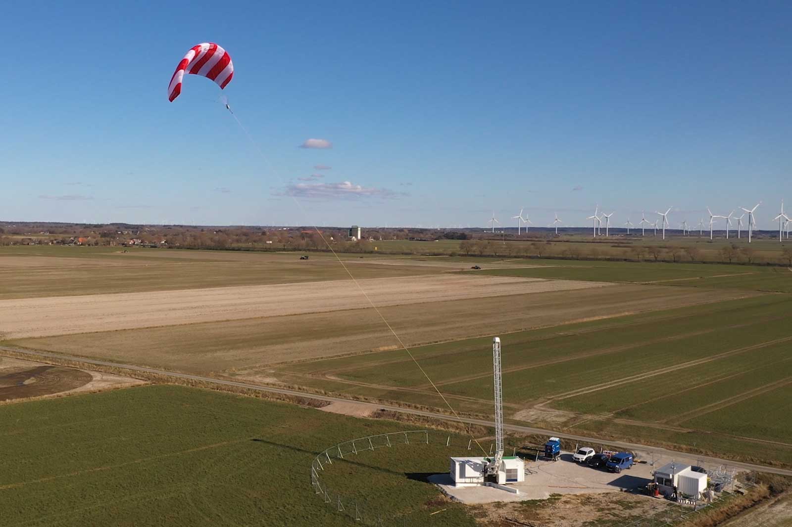 Ein rotes und weißes Fallschirmsegel schwebt über einem Feld neben Windkraftanlagen und technischen Einrichtungen.