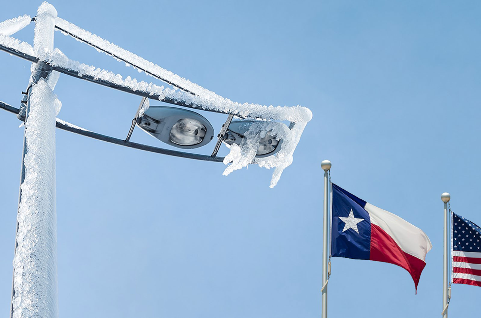 Icy light fixtures and waving flags under a clear sky, showcasing the Texas and US flags.