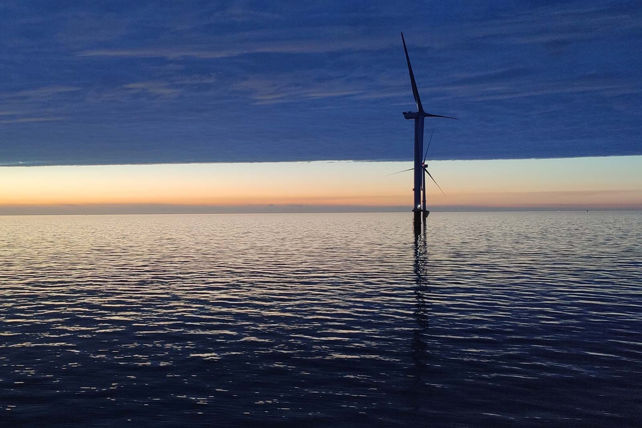 Ein Windrad steht auf einem Stützpfeiler im Wasser, während der Himmel in Lila- und Orangetönen leuchtet.