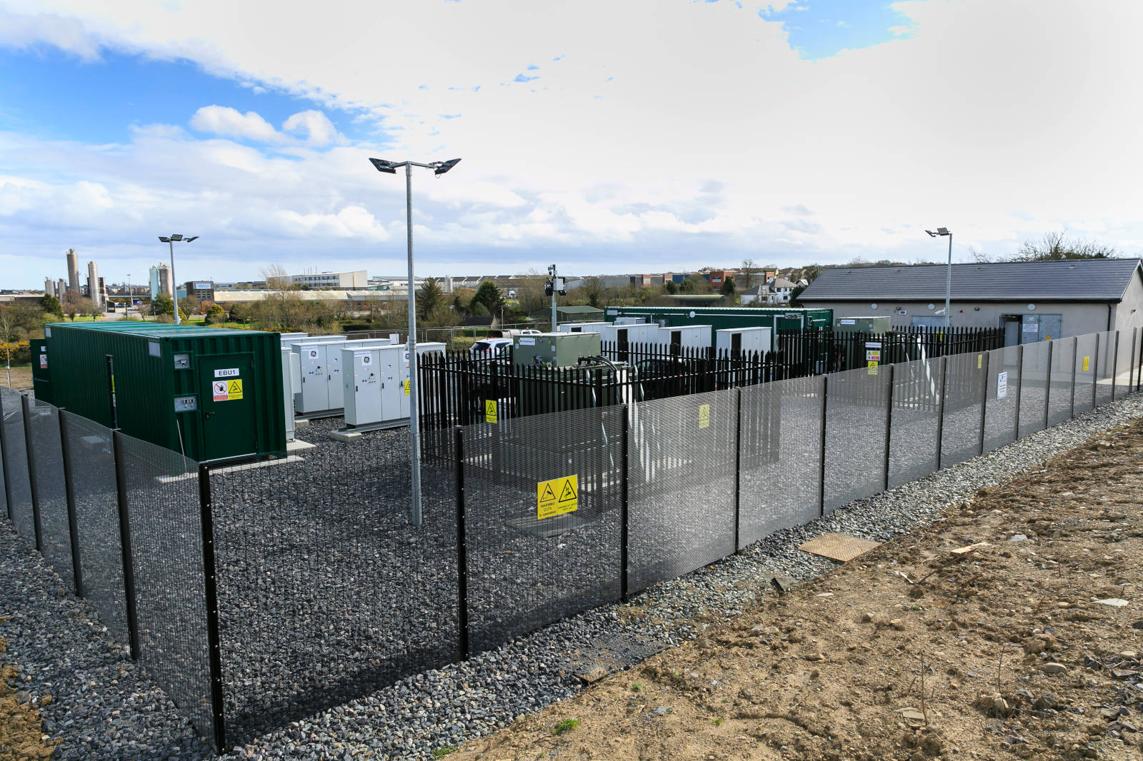 Ein eingezäunter Energiespeicherstandort mit grünen Containern und Geräten vor einem bewölkten Himmel.
