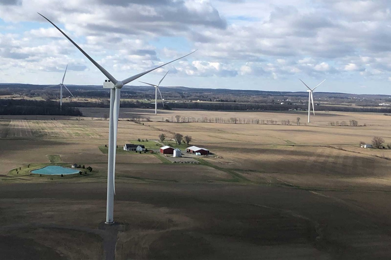 Ein weitläufiges Feld mit Windkraftanlagen und einem Bauernhof. Wolken dominieren den Himmel.