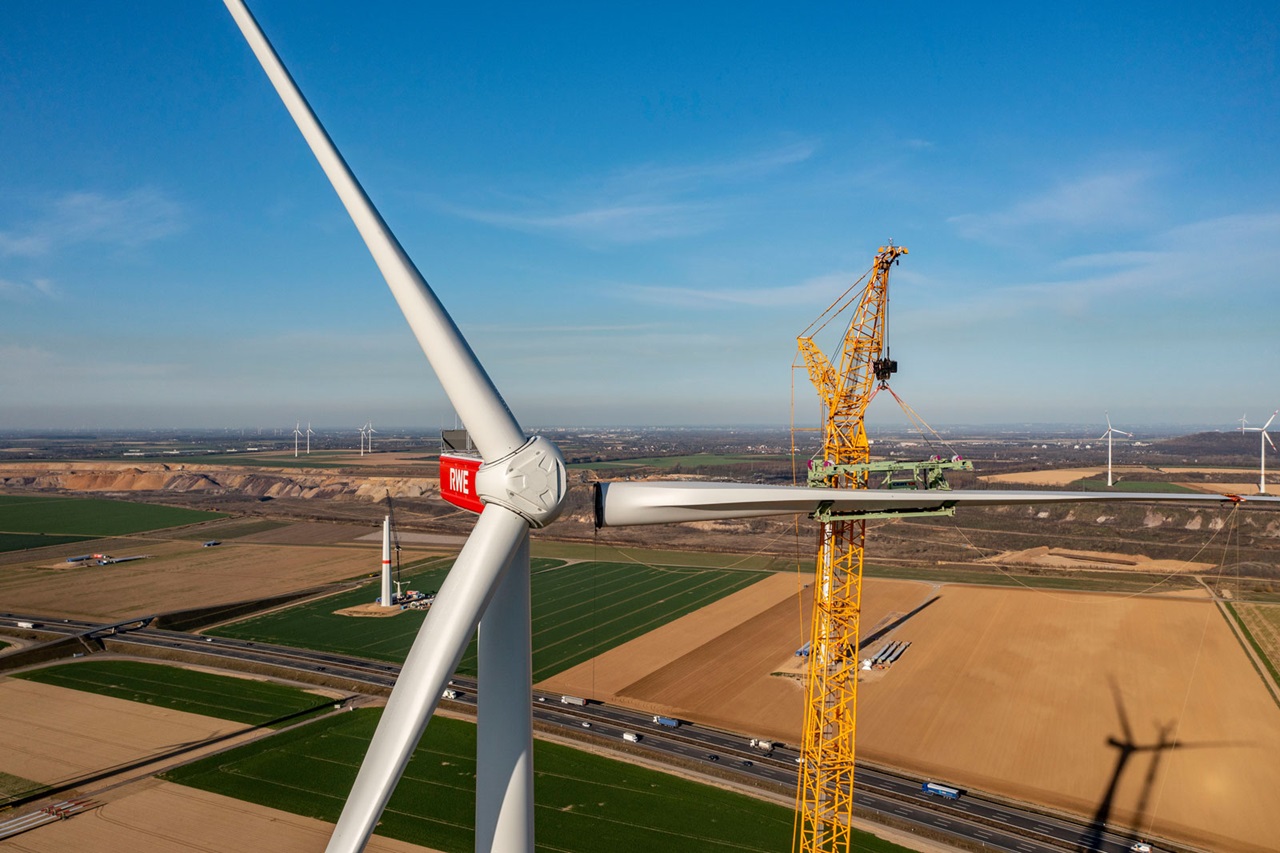 Ein Windkraftwerk mit einem Kran, der an einer Turbine arbeitet. Weite Felder und andere Windräder sind im Hintergrund sichtbar.