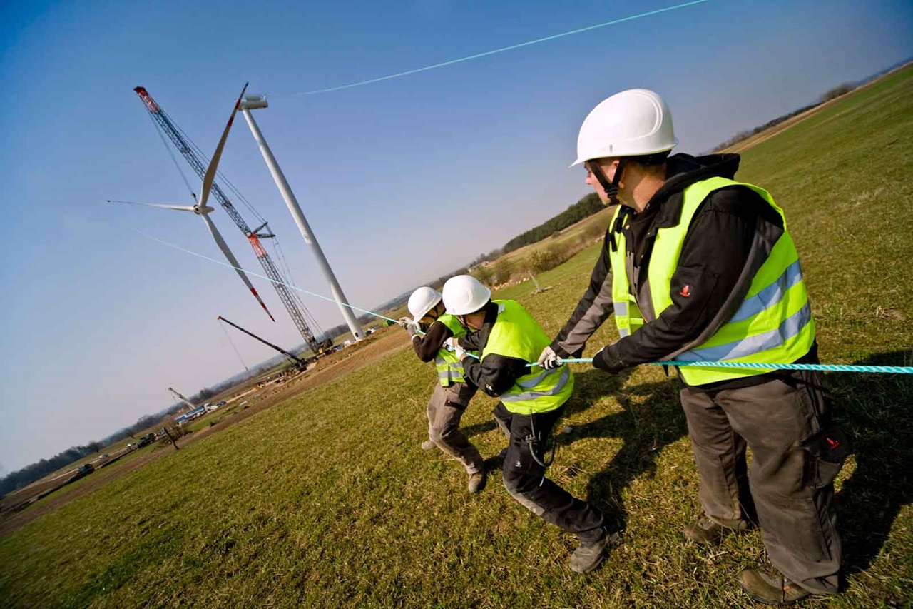 Three construction workers pull on a rope to install a wind turbine. A crane is visible in the background.