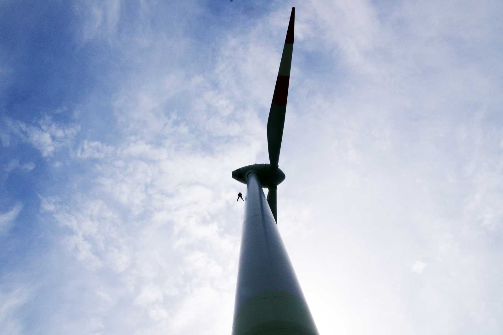 Eine Person klettert an einem hohen Windrad auf. Der Himmel ist blau mit einigen Wolken.