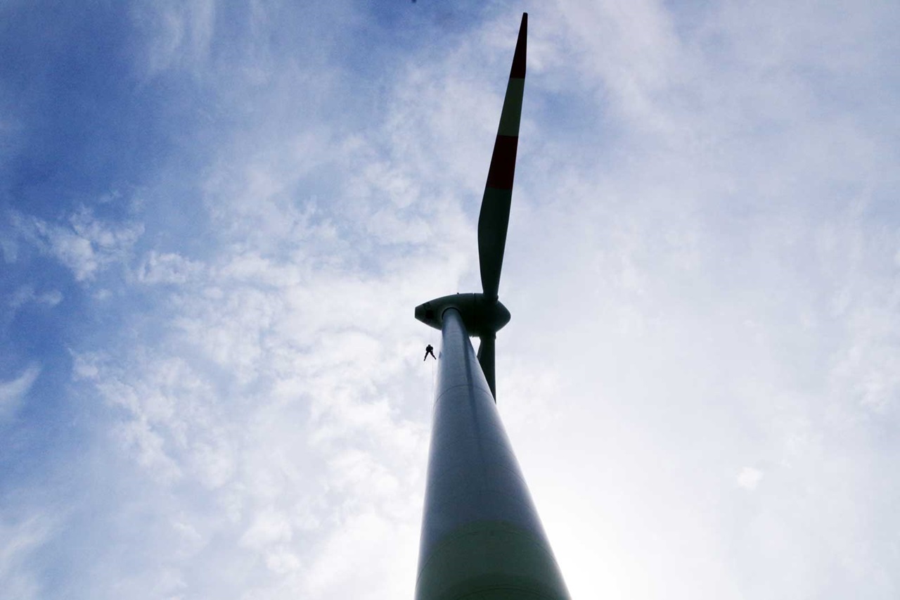 A person is climbing a tall wind turbine. The sky is blue with some clouds.