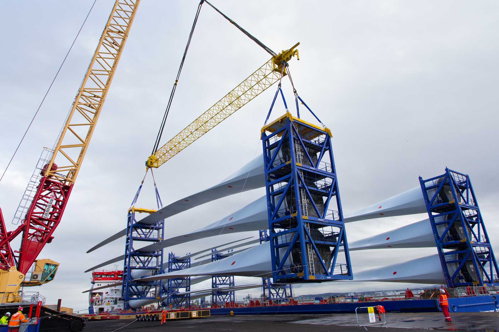 A crane lifting large wind turbine blades from a storage unit at a construction site under a cloudy sky.