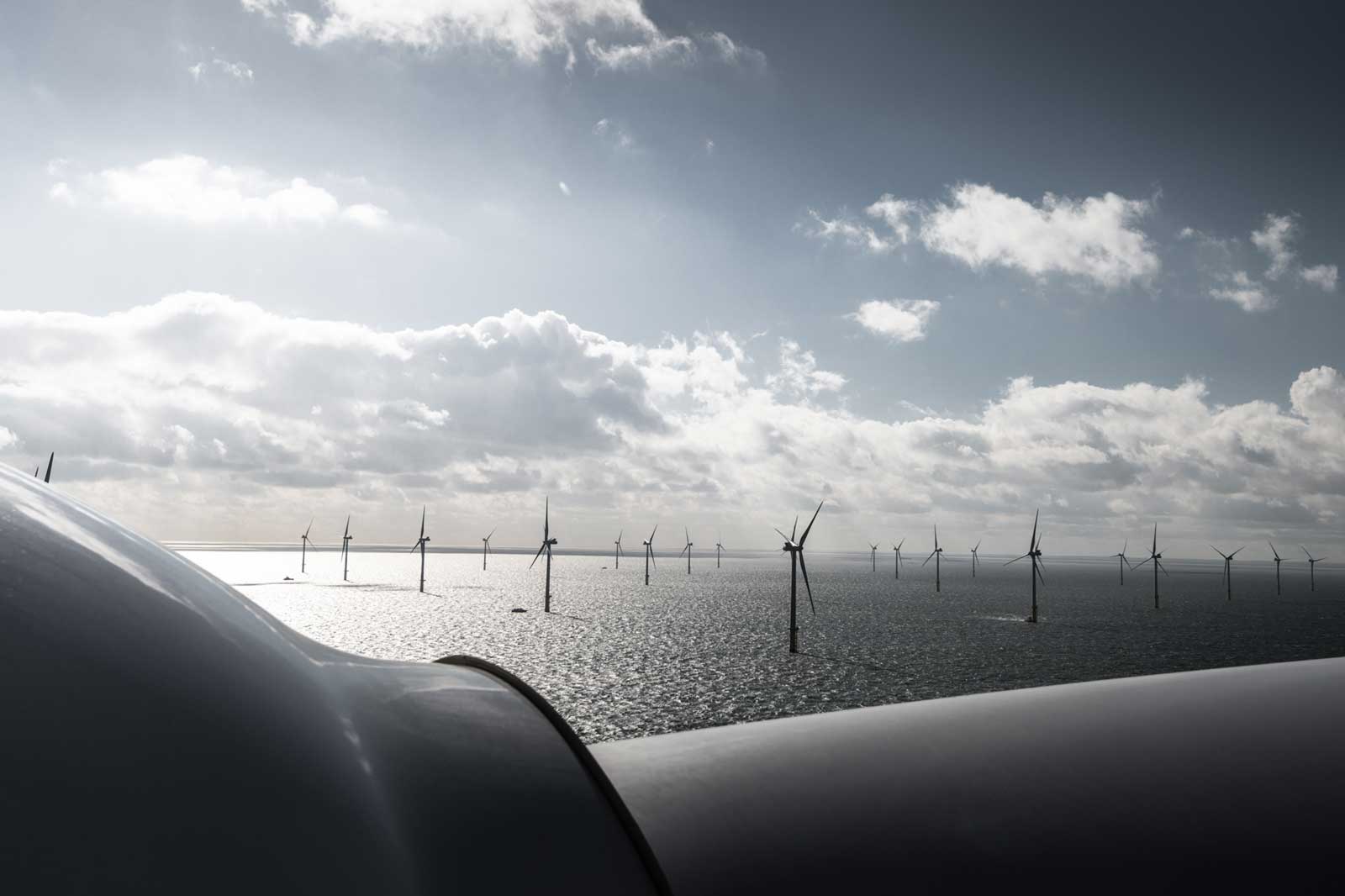 A view of offshore wind turbines with a cloudy sky above, reflecting sunlight on the water's surface.