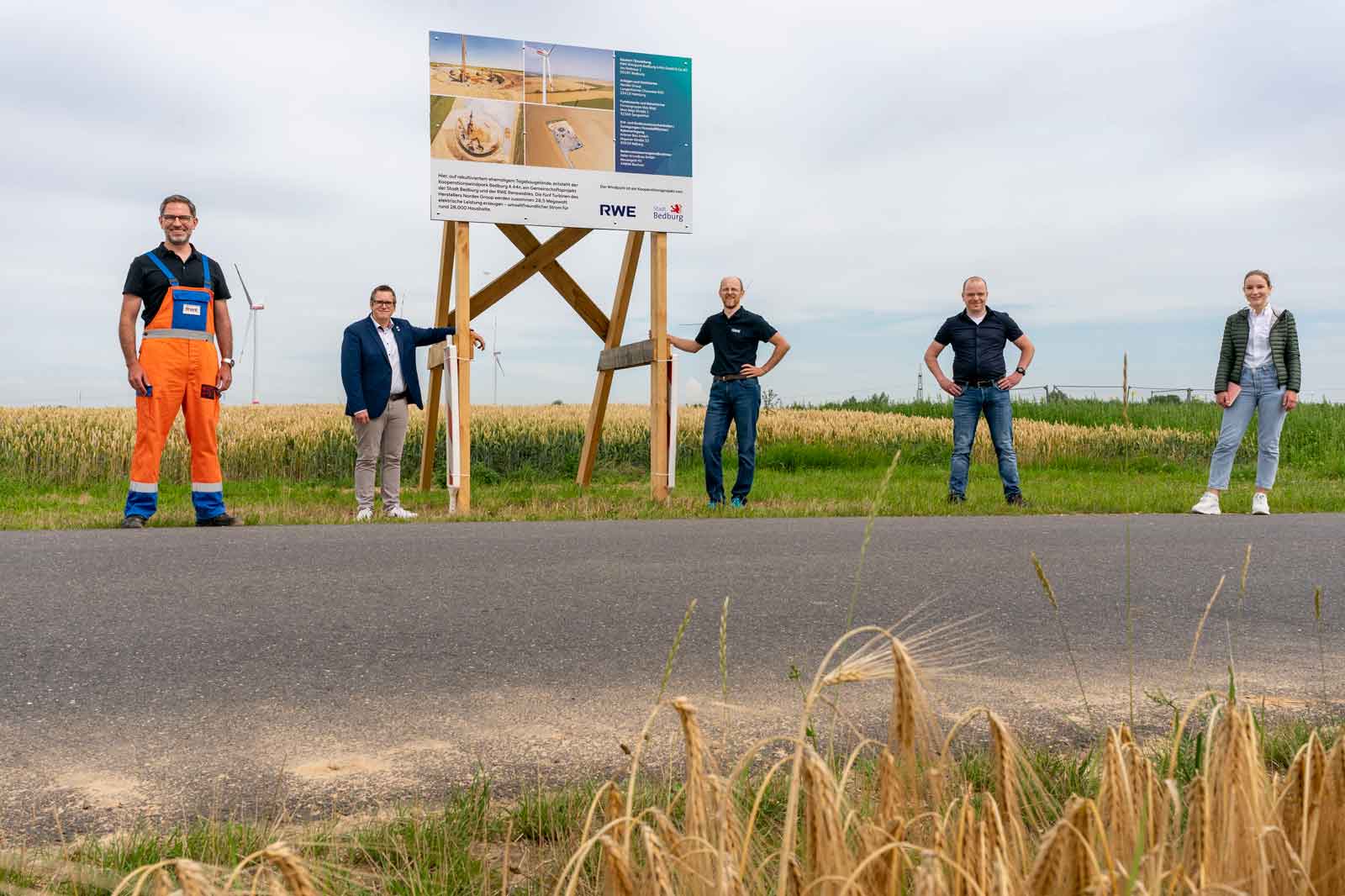 Fünf Personen stehen neben einem Schild auf einem Feld, mit Windkraftanlagen im Hintergrund sichtbar.