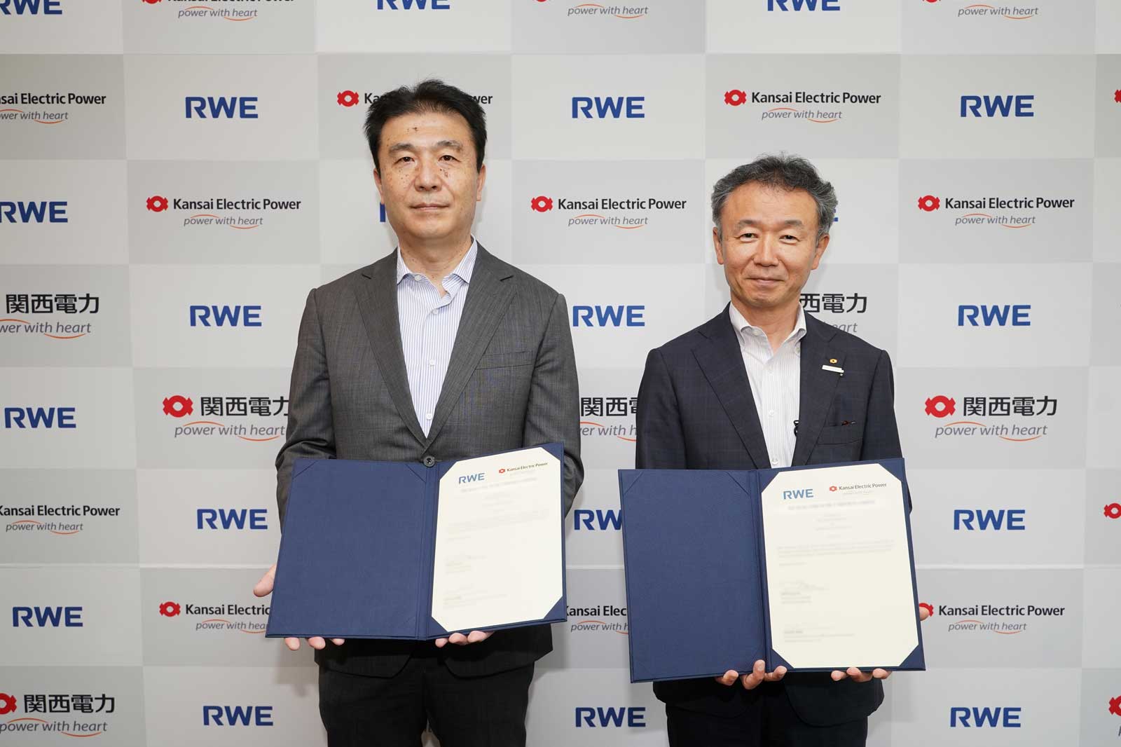 Zwei Personen halten Dokumente vor einer Wand mit Logos von Kansai Electric Power und RWE.
