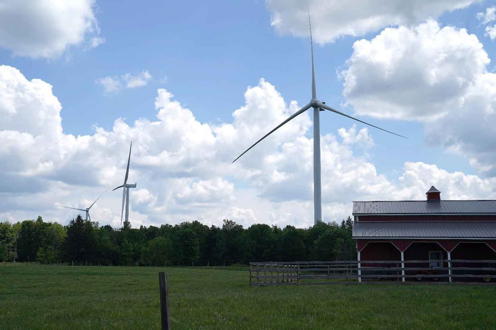 Two wind turbines stand beside a red barn on a green field, surrounded by trees under a cloudy blue sky.