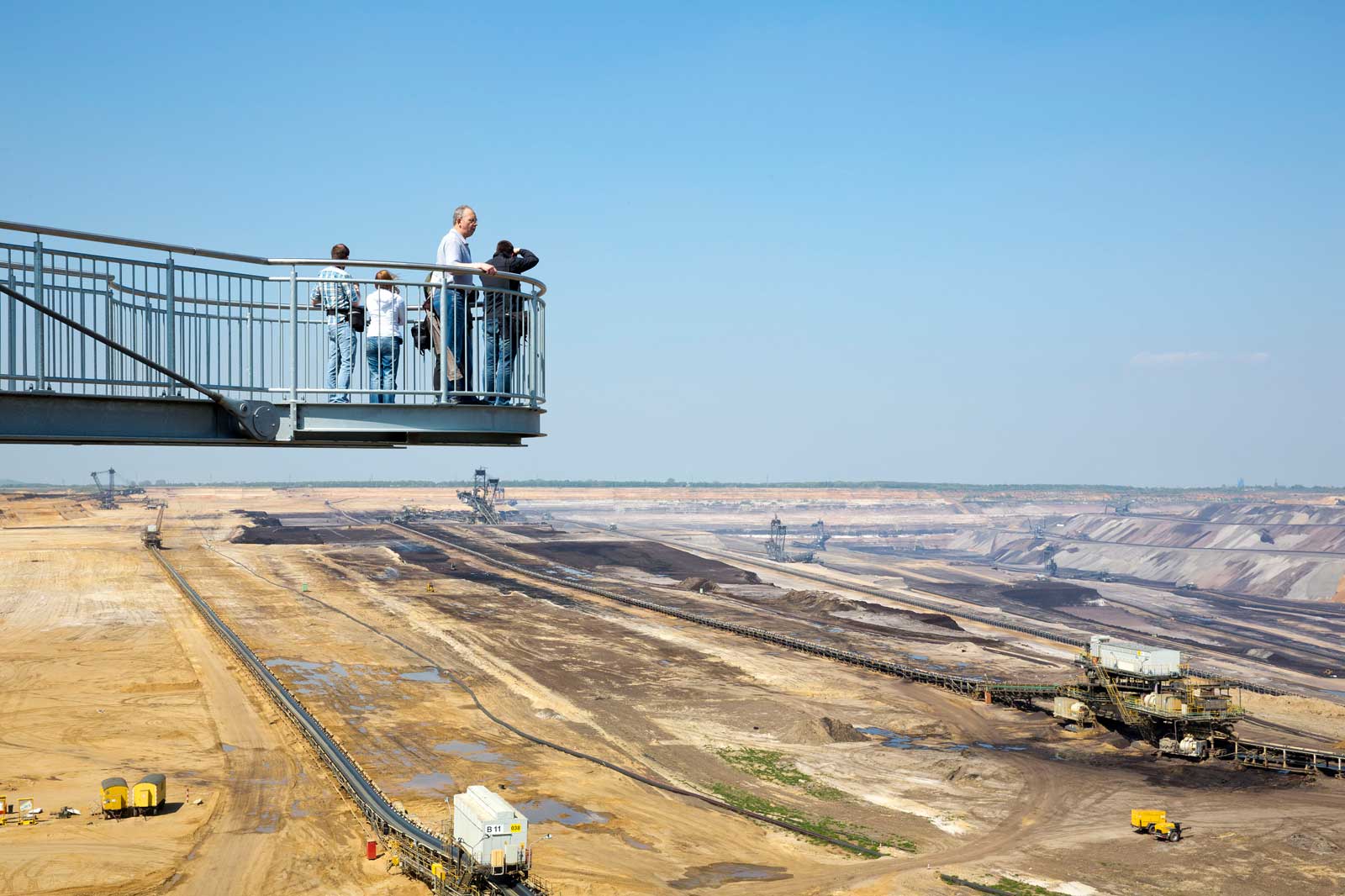 Besucher stehen auf einer Aussichtsplattform mit Blick auf eine große Bergbaustelle mit Maschinen und blauem Himmel.