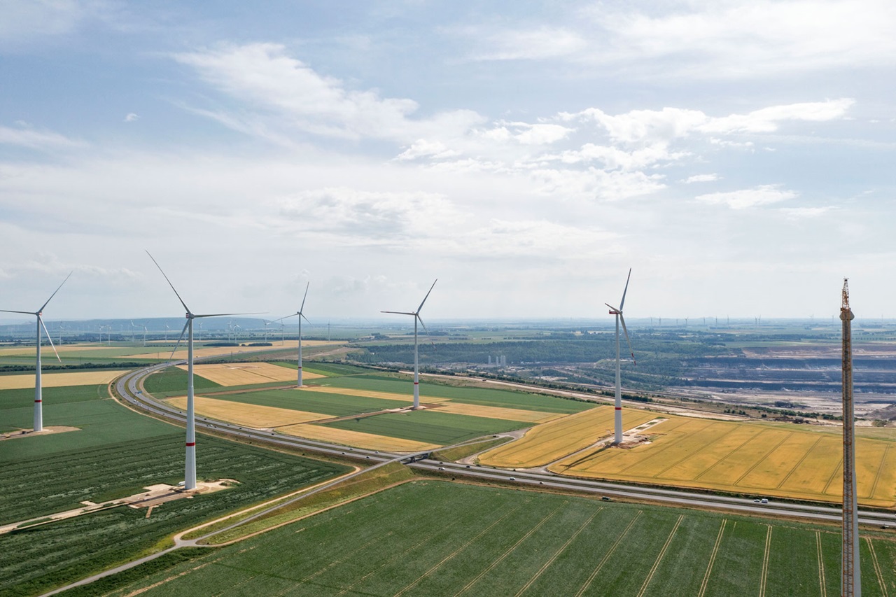 Aerial view of a wind farm with several wind turbines on green fields and a motorway in the foreground.