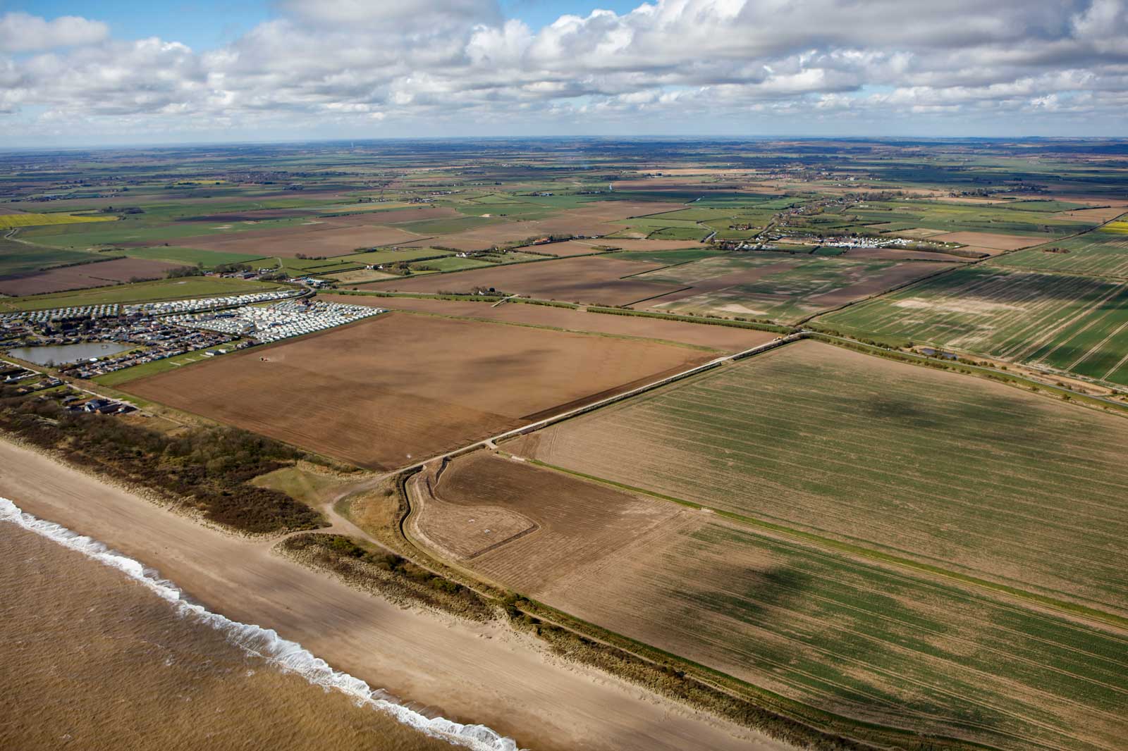 Aerial view of diverse farmland, beach, and a caravan park near the coastline under a partly cloudy sky.