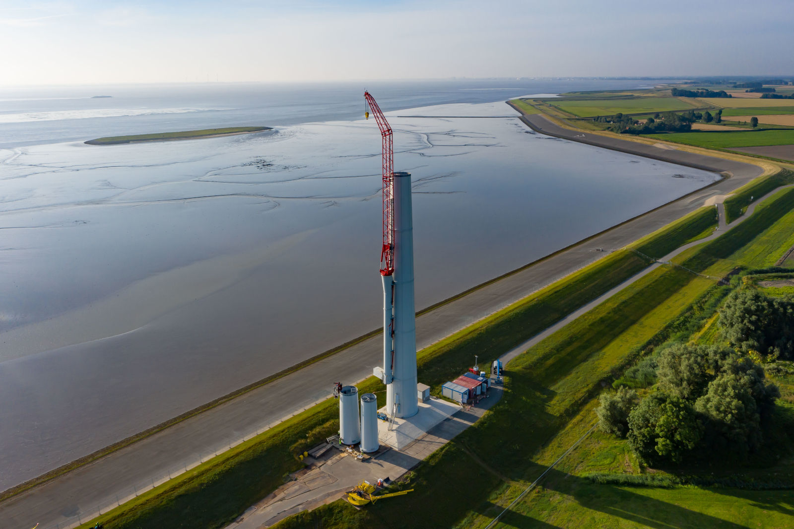 A wind turbine tower with a crane, situated by a calm water body, surrounded by meadows and fields.