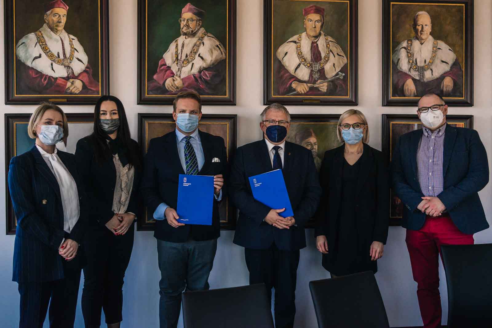 Group photo in a room with historical portraits. Various individuals holding documents in blue folders.