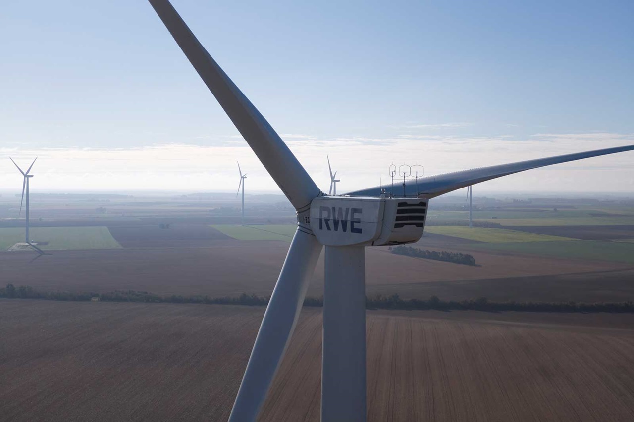 Nahaufnahme eines Windrads mit dem Schriftzug RWE, im Hintergrund weitere Windkraftanlagen und ein landwirtschaftliches Feld.