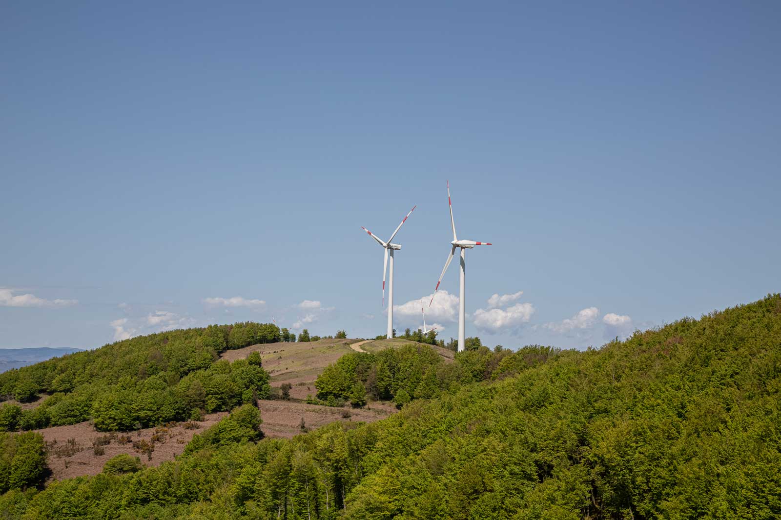 Zwei Windkraftanlagen stehen auf einem Hügel in einer bewaldeten Landschaft unter einem blauen Himmel.