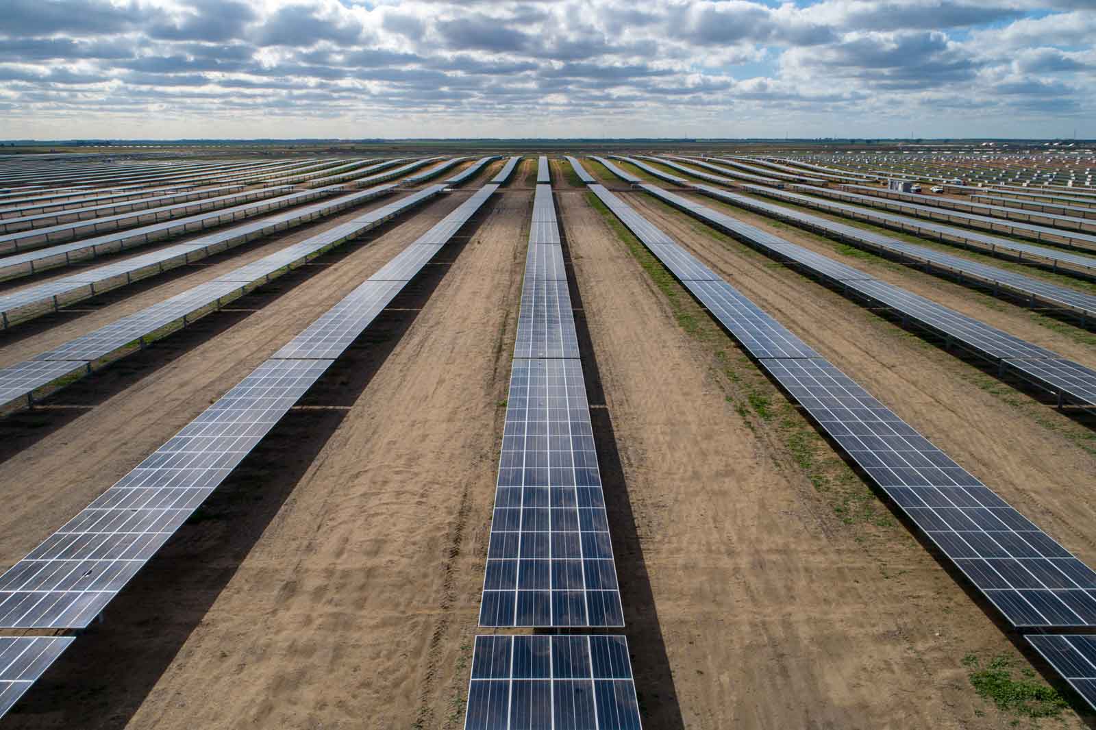 An expansive solar farm with numerous rows of solar panels under a partly cloudy sky, surrounded by a lush landscape.