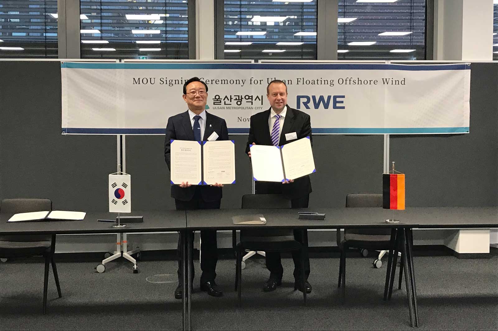 Two men hold signed documents during an MOU signing ceremony, with flags in the background.