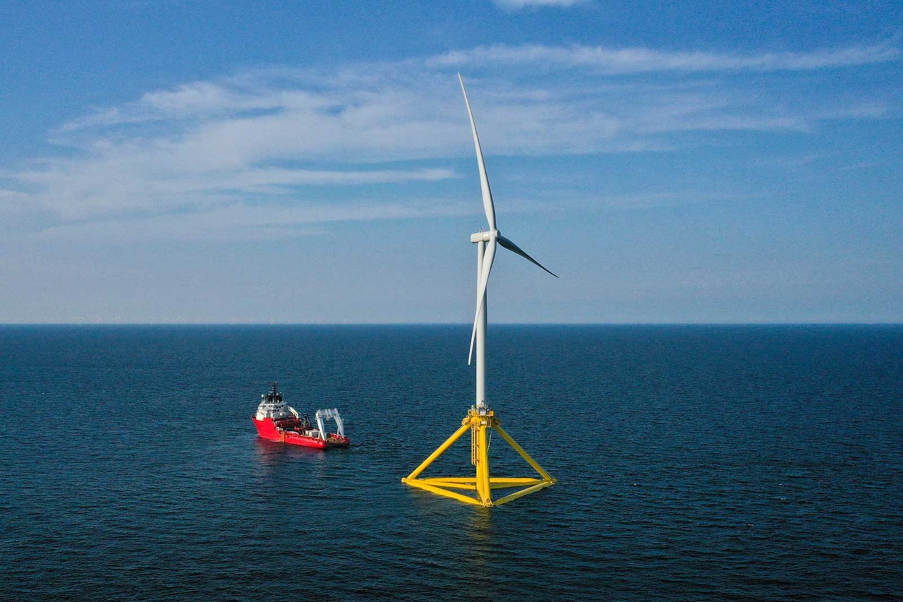 Ein Windkraftwerk steht im Meer, flankiert von einem Schiff. Der Himmel ist blau mit leichten Wolken.