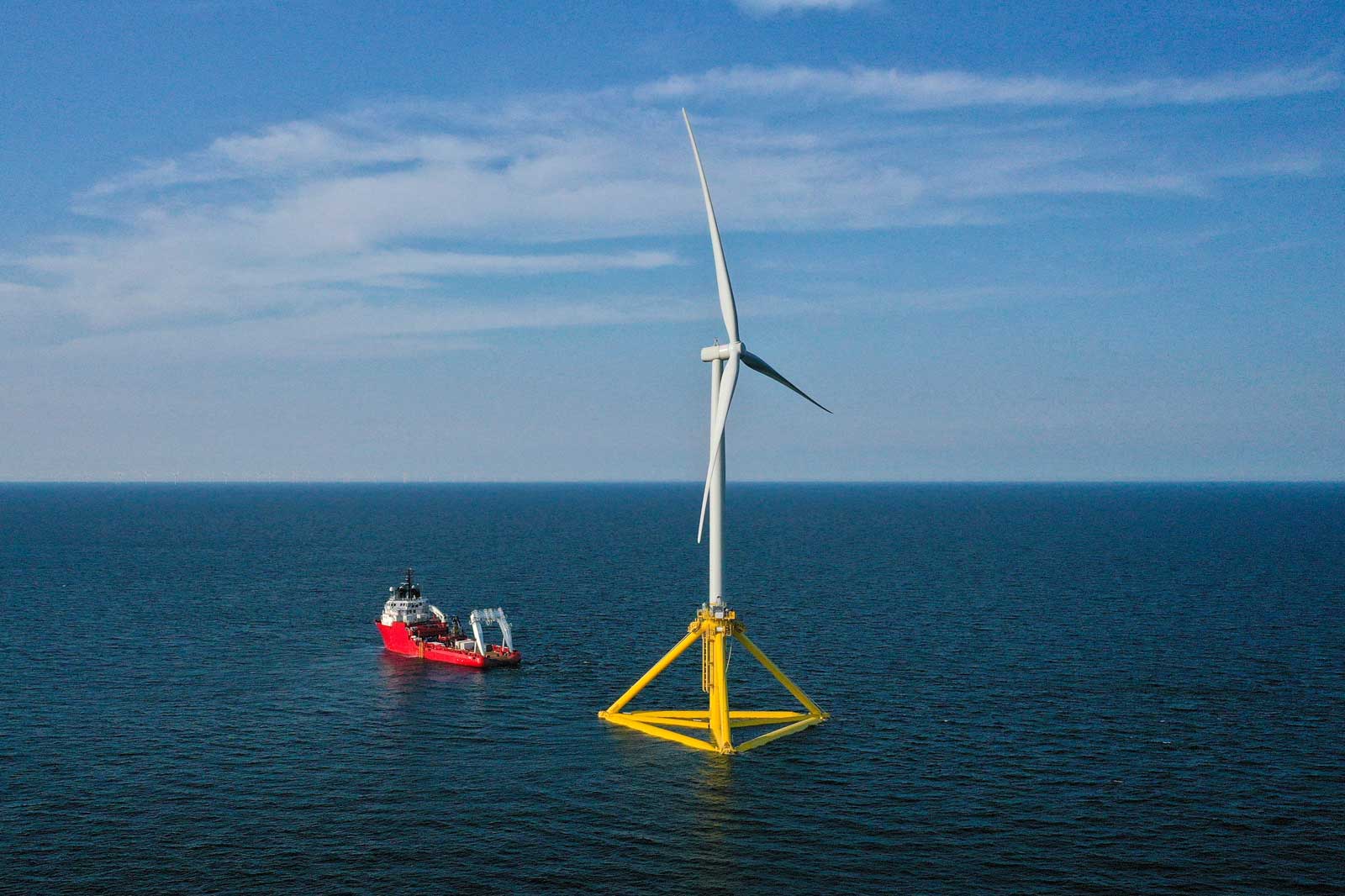 A wind turbine stands in the sea, accompanied by a vessel. The sky is blue with light clouds.