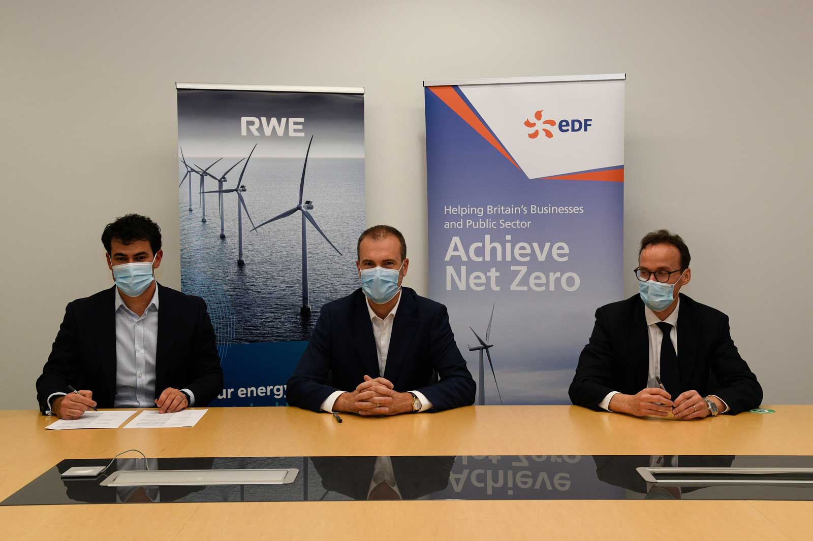 Three individuals sit at a table. Background features RWE and EDF banners representing wind energy initiatives.