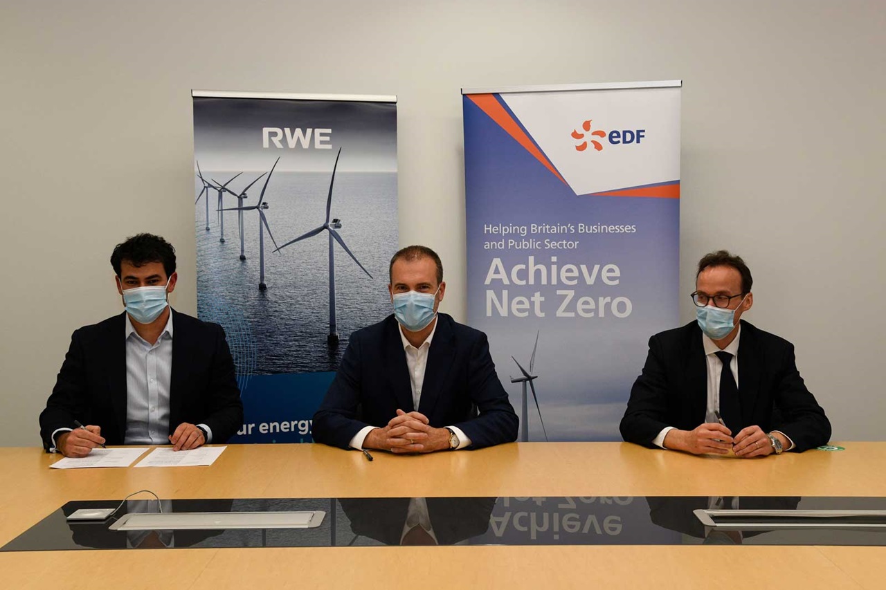 Drei Personen sitzen an einem Tisch. Im Hintergrund sind Banner von RWE und EDF zu sehen, die Windkraft symbolisieren.