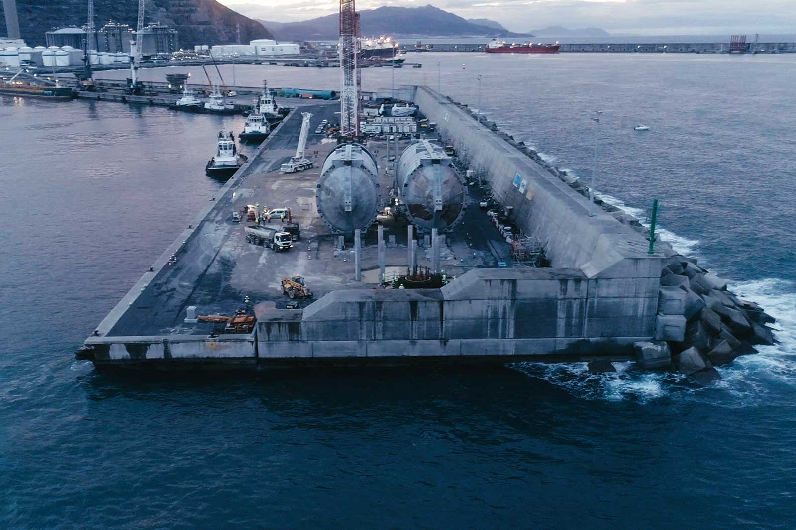 An aerial view of a dock with large industrial containers, construction vehicles, and boats in a harbour at dusk.