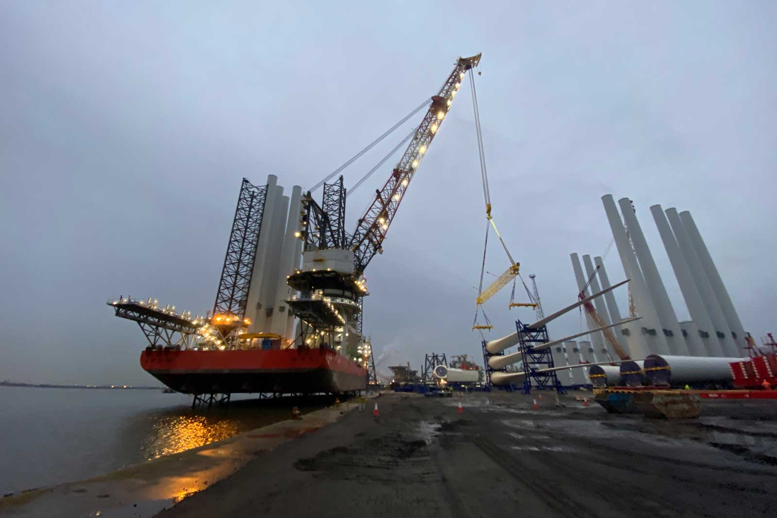 A large crane on a vessel lifts a structure at a construction site near water, with wind turbine components in the background.