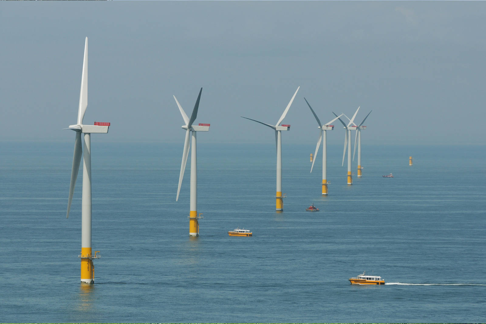 A sea view featuring multiple wind turbines and small boats in the foreground.