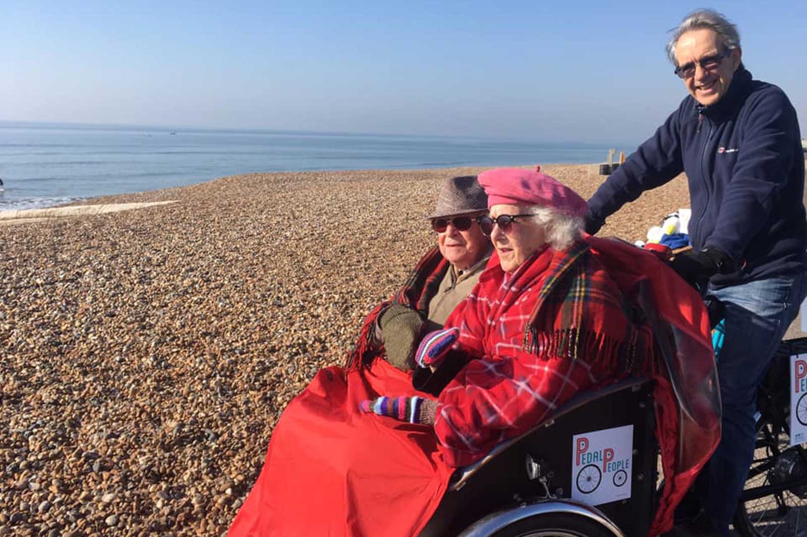 An older woman in a wheelchair covered with a red blanket, at the beach, pushed by a man. The sea is calm with a clear sky.