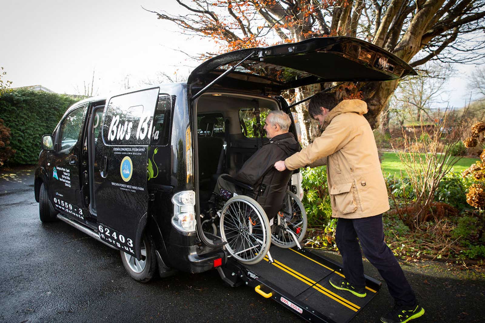 A wheelchair user is being assisted into a black accessible vehicle with a ramp in a garden setting.