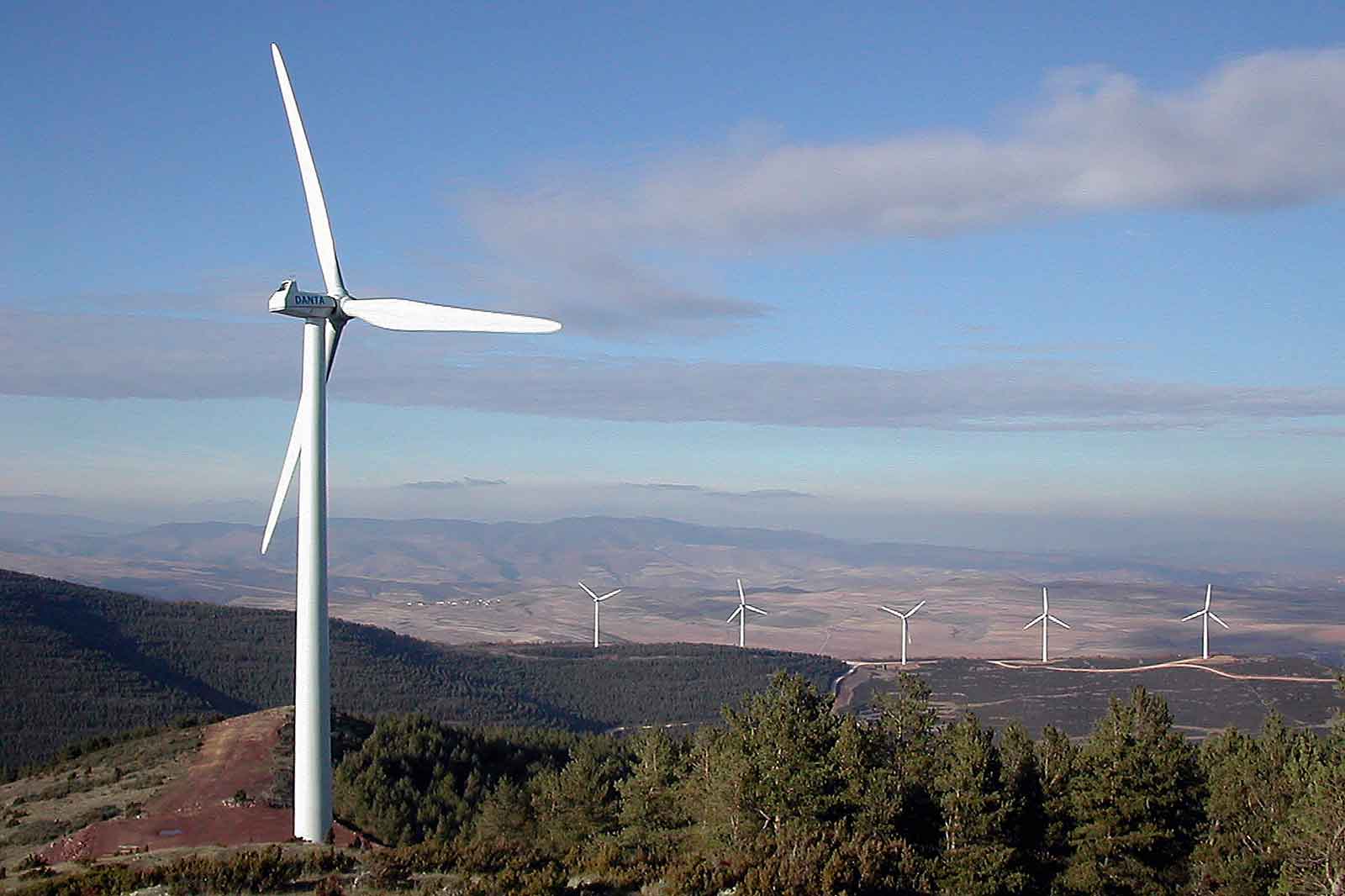 Ein einzelner Windkraftanlage auf einem Hügel, umgeben von Bäumen und weiteren Windkraftanlagen im Hintergrund.