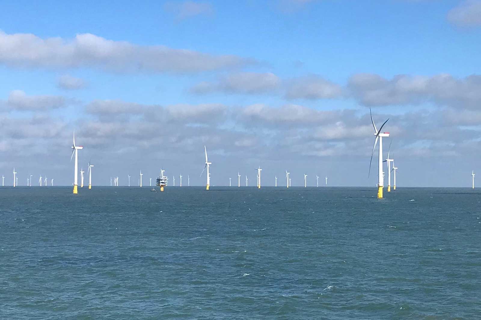 A view of offshore wind turbines in the sea under a partly cloudy sky, with a service vessel nearby.