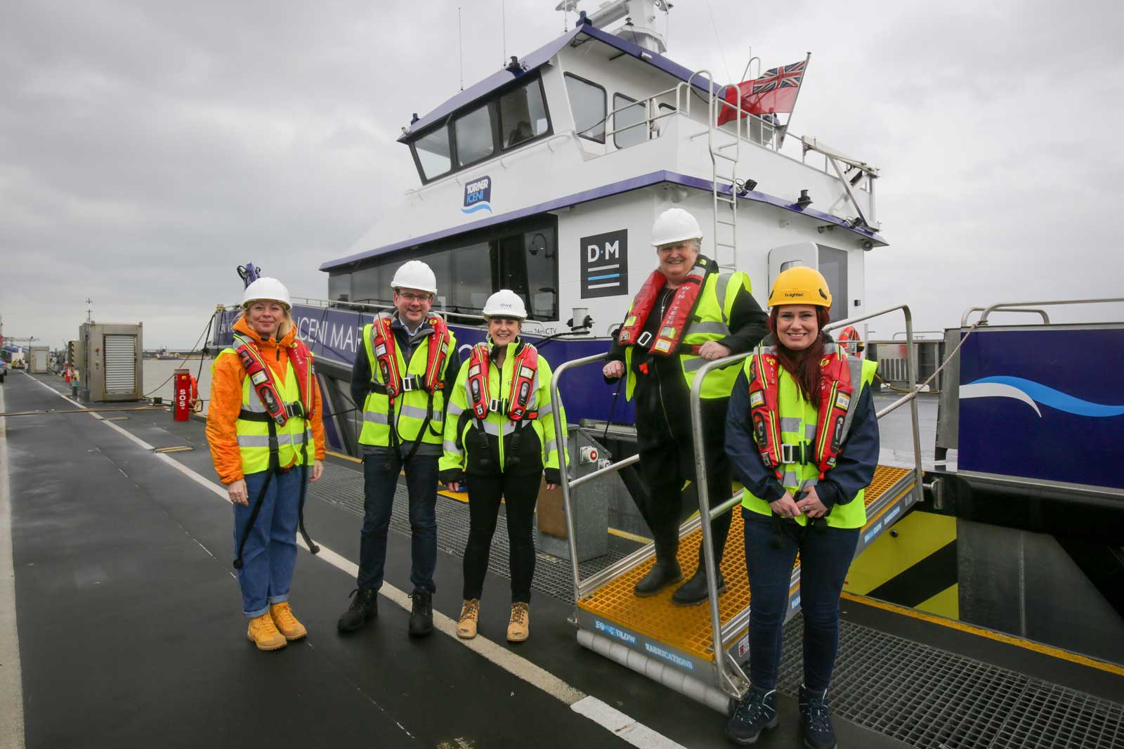 Five individuals in safety vests and helmets stand near a vessel at a port on a cloudy day.