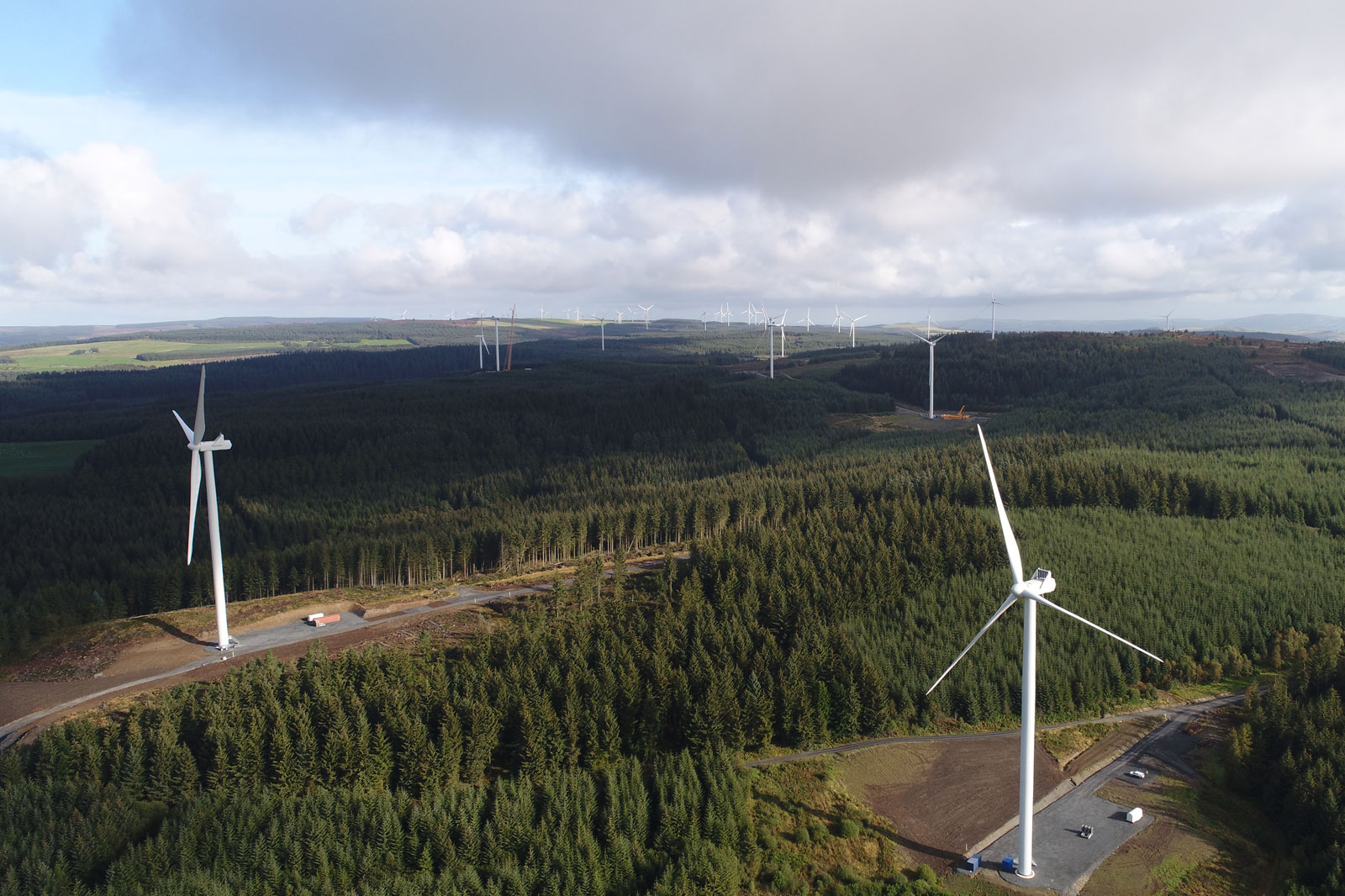 Aerial view of a wind farm with several wind turbines surrounded by dense forests and hilly terrain under a cloudy sky.