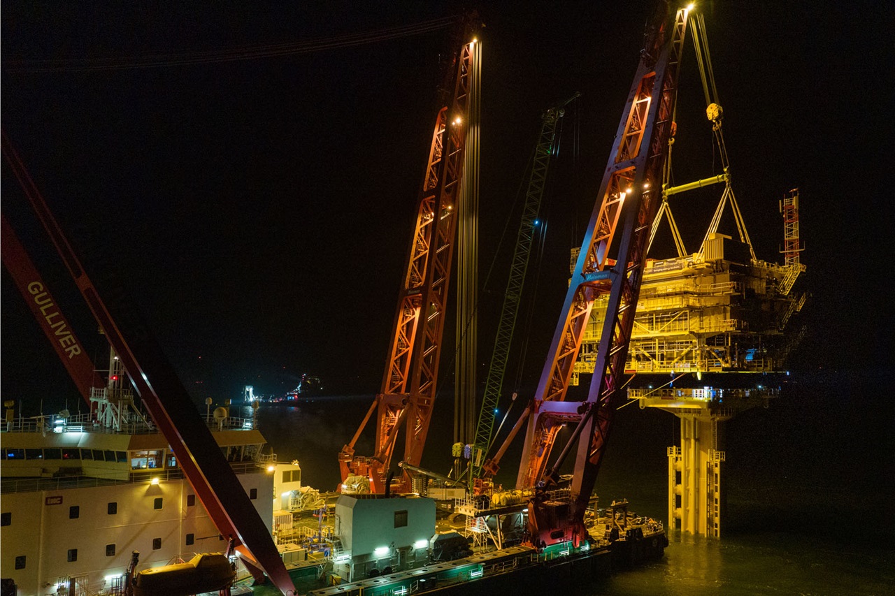An offshore platform is being lifted at night by a crane from a ship. The crane and ship are brightly lit.