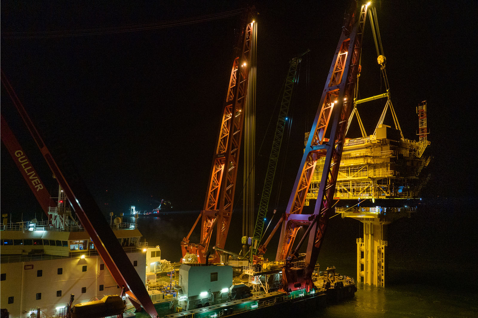 An offshore platform is being lifted at night by a crane from a ship. The crane and ship are brightly lit.